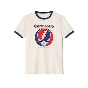 May include: Cream-colored ringer t-shirt featuring the Grateful Dead logo. The logo is a skull with a lightning bolt, set within a red and blue circle. The text "Grateful Dead" is above the logo in red script. The shirt has navy blue trim.
