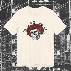 May include: Cream-colored t-shirt featuring a detailed black and white skull design, adorned with a wreath of red roses. The graphic tee has a classic crew neck and short sleeves, suitable for casual wear.