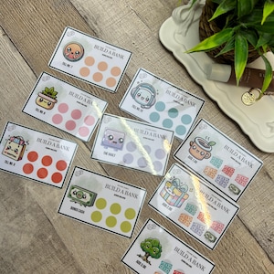 May include: A collection of Build a Bank cards with various designs and illustrations. Each card features a different theme, such as a cute face, a shopping bag, or a gift box, along with a set of circles for tracking savings. The cards are on a wooden surface.