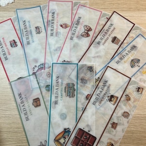 May include: A collection of translucent bookmarks with various designs and text. Each bookmark features the words "BUILD A BANK" and a different theme, such as "HELLO DOCTOR," "BATHROOM BREAK," and "SELFCARE DAY." The bookmarks have colorful illustrations.