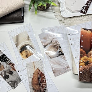 Può includere: Una collezione di inserti in plastica trasparente per sfide di risparmio per raccoglitori, ognuno con una fotografia stagionale unica, tra cui caffè, zucche e foglie autunnali, con il testo stampato COZY CHALLENGE e 25 ENVELOPE.