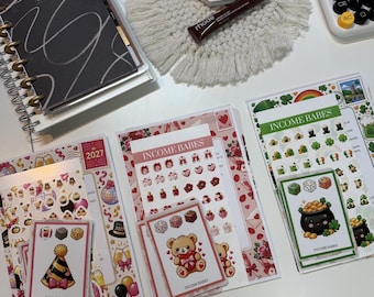 May include: A black spiral-bound planner, a white Fenty Beauty compact, a brown Rhode lip treatment, a white calculator, and three sets of Income Babes sticker sheets featuring illustrations of party hats, teddy bears, and pots of gold.