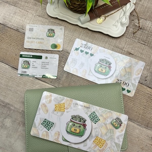 May include: An overhead shot features a green leather wallet, debit card, and Banker ID card. The cards have a cartoon jar of money graphic. Two $25.00 bills are also visible, along with a decorative tray with a plant and beauty product.