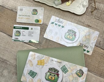 May include: An overhead shot features a green leather wallet, debit card, and Banker ID card. The cards have a cartoon jar of money graphic. Two $25.00 bills are also visible, along with a decorative tray with a plant and beauty product.
