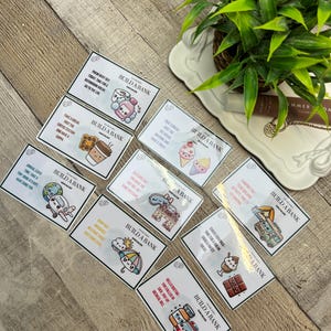 Pode incluir: Uma coleção de cartões retangulares com várias ilustrações e texto, dispostos sobre uma superfície de madeira. Cada cartão apresenta a frase "BUILD A BANK" juntamente com imagens de um avião, sorvete, café e outros itens. Uma planta em vaso está no fundo.