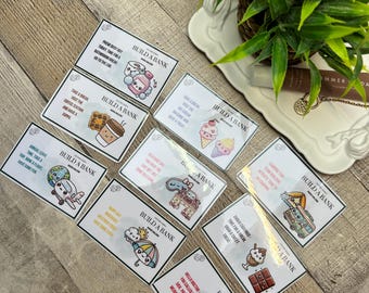 May include: A collection of rectangular cards with various illustrations and text, arranged on a wooden surface. Each card features the phrase "BUILD A BANK" along with images of an airplane, ice cream, coffee, and other items. A potted plant is in the background.
