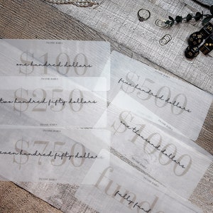 May include: A collection of translucent gift cards with varying dollar amounts, including "one hundred dollars," "two hundred fifty dollars," and "one thousand dollars." The cards are arranged on a wooden surface, with a pearl necklace, rings, and other decorative items.