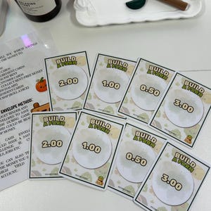 Könnte beinhalten: Eine Sammlung von "Build a Farm" Spielkarten mit unterschiedlichen Einkommenswerten, von 0,50 bis 3,00. Die Karten zeigen ein kreisförmiges Design mit einer Farm-Szene und einer kleinen Cartoon-Figur. Ebenfalls sichtbar sind Schönheitsprodukte und eine Kerze.