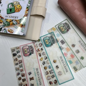 以下が含まれることがあります： 「Build a Bank Academy」のデザインが施されたベージュ色のオープンなプランナーと、いくつかの装飾的なブックマーク。ブックマークには漫画のキャラクターと、お金をテーマにしたイラストが描かれており、「All Angry Customers」と「All Happy Customers」というテキストが書かれています。