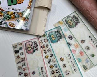 May include: An open planner with a beige cover and a "Build a Bank Academy" design, alongside several decorative bookmarks. The bookmarks feature cartoon characters and money-themed illustrations, with text that reads "All Angry Customers" and "All Happy Customers."