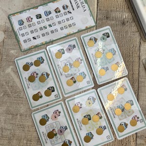 Puede incluir: Un conjunto de tarjetas de desafío de ahorro "Build a Bank". Las tarjetas presentan ilustraciones de personajes de dibujos animados, dinero y dados. Cada tarjeta tiene una moneda de oro adjunta. El texto "Build a Bank" está impreso en las tarjetas.