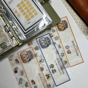 Puede incluir: Una colección de pegatinas y accesorios de planificación financiera. Las pegatinas presentan ilustraciones de botes de dinero y personajes lindos. La frase "Build a Bank" es visible en las pegatinas. Una agenda verde y una cartera marrón también están en la imagen.