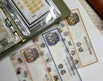 May include: A collection of financial planning stickers and accessories. The stickers feature illustrations of money jars and cute characters. The phrase "Build a Bank" is visible on the stickers. A green planner and a brown wallet are also in the image.