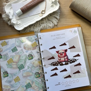 May include: A yellow planner with pages featuring money and a pig graphic. A tube of Summer Fridays lotion and a small gold charm are on a white tray. A beige clutch is also visible.