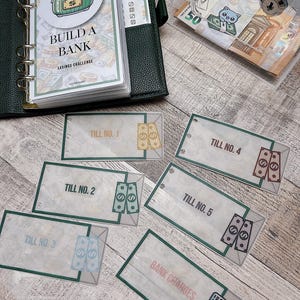 May include: A green binder with the words "Build a Bank Savings Challenge" on the cover. There are several clear plastic envelopes with the words "Till No." and a dollar sign on each. There is also a clear plastic box with a lock and a 50 euro note inside.