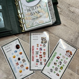 May include: A green binder with gold rings and a savings challenge insert that says "Build a Bank." Three rectangular cards feature the text "Build a Bank" and various animal and emoji icons. The cards are labeled "Extra Customers," "Angry Customers," and "Happy Customers."