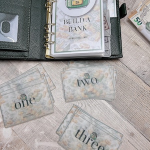 Puede incluir: Una carpeta verde con un desafío de ahorro titulado "Construir un banco" con una bolsa de dinero de dibujos animados en la portada. Hay tres tarjetas con los números "uno", "dos" y "tres" en ellas. Las tarjetas tienen un patrón de bolsa de dinero.
