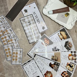 May include: A collection of autumn-themed cards and stickers with various designs, including plaid patterns, leaves, and pumpkins. Some cards feature the word "AUTUMN" and other text. A white tray with a tube of "SUMMER FRIDAYS" product is also present.