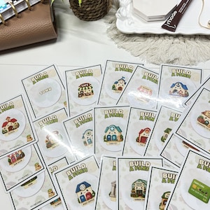 May include: A collection of Build a Farm game cards featuring various illustrated buildings like a cafe, school, and post office, alongside a ring, a Fenty Beauty compact, and a Rhode lip tint tube arranged on a white surface with a plant.