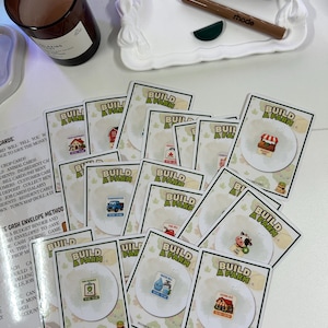 以下が含まれることがあります： 「Build a Farm」ゲームカード、ガラス瓶に入った茶色のキャンドル、2つのリップ製品が入った白いトレイを真上から見た写真。カードには農場をテーマにしたイラストとテキストが描かれています。