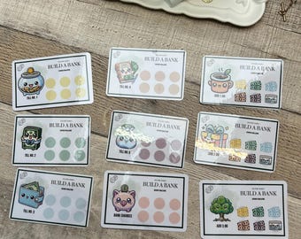 May include: Nine Build a Bank cards with cute illustrations and text. Each card features a different design, including a piggy bank, a wallet, and a gift box. The cards have spaces for tracking savings and are set against a light wooden surface.