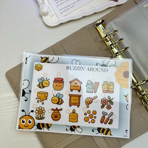 Puede incluir: Una agenda con anillos dorados y un bolsillo transparente que contiene pegatinas con temática de abejas. Las pegatinas presentan abejas de dibujos animados, tarros de miel y el texto "BUZZIN' AROUND". Una bandeja blanca contiene un tubo de bálsamo "SUMMER FRIDAYS" y una nota.
