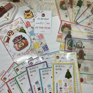 Puede incluir: Una colección de suministros de presupuesto con temática navideña. Incluye sobres de plástico transparente con diseños festivos, tarjetas con el texto "it's the most wonderful time of the year" y tarjetas "Build a Bank". La paleta de colores incluye rojo, verde y dorado.