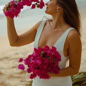 May include: A woman in a white dress holds vibrant pink bougainvillea flowers on a sandy beach. The flowers are in full bloom, with a rich, deep pink colour. The woman is smiling, enjoying the flowers.