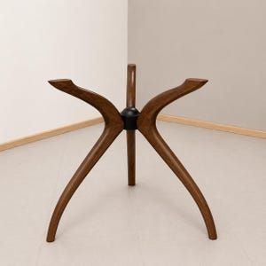 May include: A wooden table base with a unique, modern design. The base features three curved legs that extend outward from a central black connector. The legs are a warm brown color, and the design is ideal for a round table top.