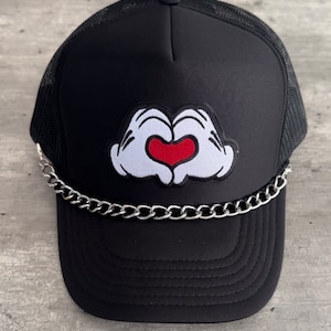 May include: Black trucker hat with a white and red heart-shaped hand gesture patch and a silver chain around the brim.
