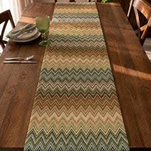 May include: A long rectangular table runner featuring a repeating chevron pattern in shades of brown, tan, olive green, and cream. The fabric displays a textured weave with horizontal zig-zag lines running across the entire length of the runner.