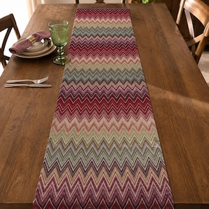 Könnte beinhalten: Ein rechteckiger Tischläufer mit einem sich wiederholenden Zickzackmuster in einer lebendigen Farbpalette aus Rot, Burgunder, Rosa, Beige und Salbeigrün. Das Textil zeigt ein durchgehendes Zickzack-Design, das sich über die Länge erstreckt.
