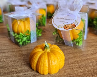 May include: A set of small, clear boxes with white ribbon tied around them. Each box contains a small, yellow pumpkin-shaped candle. The boxes are arranged on a wooden surface. A larger, yellow pumpkin-shaped candle is in the foreground. The boxes have a sticker that says "Bridal Shower 07.23.2023".