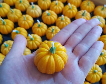 May include: A hand holding a small, yellow pumpkin-shaped candle. The candle is surrounded by many other similar candles, all arranged in a wooden tray.