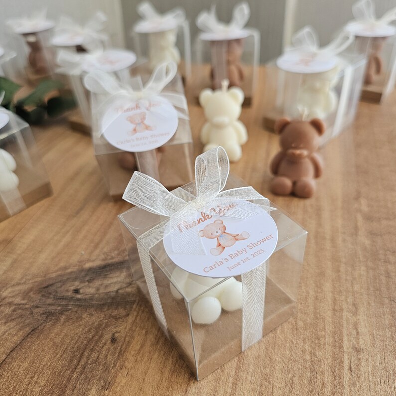 May include: A set of small, clear boxes with white and brown teddy bear candles inside. Each box has a white circle sticker with a brown teddy bear and the text "Thank You Carla's Baby Shower June 18, 2023".