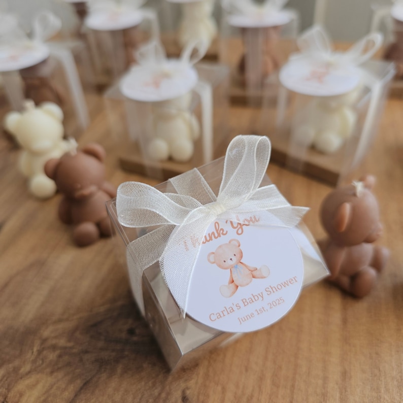 May include: A small clear box with a white ribbon and a thank you tag. The tag has a brown teddy bear with a blue bow tie and the text "Carla's Baby Shower June 1st, 2025".