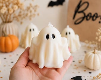 May include: A white ghost-shaped candle with two black oval eyes and a small wick at the top. The candle has a draped, flowing appearance resembling a sheet. It is held in a palm, with other similar ghost candles and small pumpkins nearby.