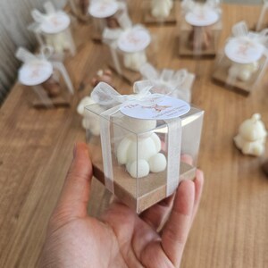May include: A small white bear figurine in a clear plastic box with a brown base. The box is tied with a white ribbon and has a small white circle label with the text "Tika" and "Carter's Baby Shower" on it.
