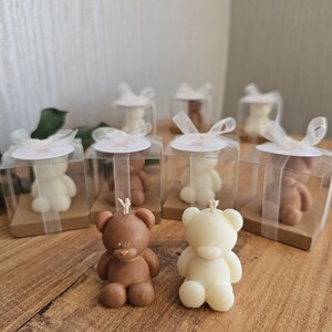 Puede incluir: Un conjunto de peque&ntilde;as velas de oso de peluche marrones y blancas en cajas transparentes con tapas marrones y cintas blancas. Las velas est&aacute;n dispuestas sobre una superficie de madera.