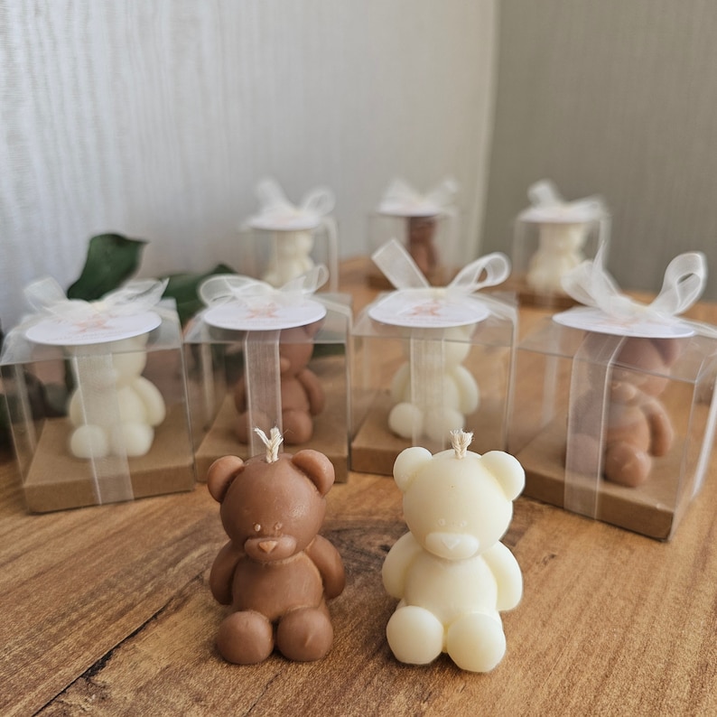 May include: A set of small, white and brown teddy bear candles in clear boxes with brown lids and white ribbons. The candles are arranged on a wooden surface.