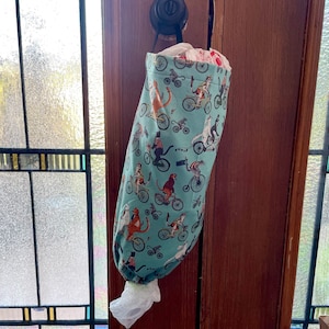 May include: A teal fabric bag with a drawstring closure, hanging on a door. The bag is covered in a pattern of cats riding bicycles.