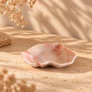 May include: A small, shallow, organic-shaped dish featuring a white base with swirling pink and peach marble-like patterns. The edges are uneven and wavy, mimicking the natural form of a shell or a smooth stone, with a polished, glossy finish.