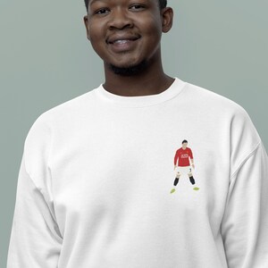 May include: A white sweatshirt with a red and white illustration of a soccer player wearing a red jersey with the number 7 on it. The player is wearing black shorts and yellow cleats.