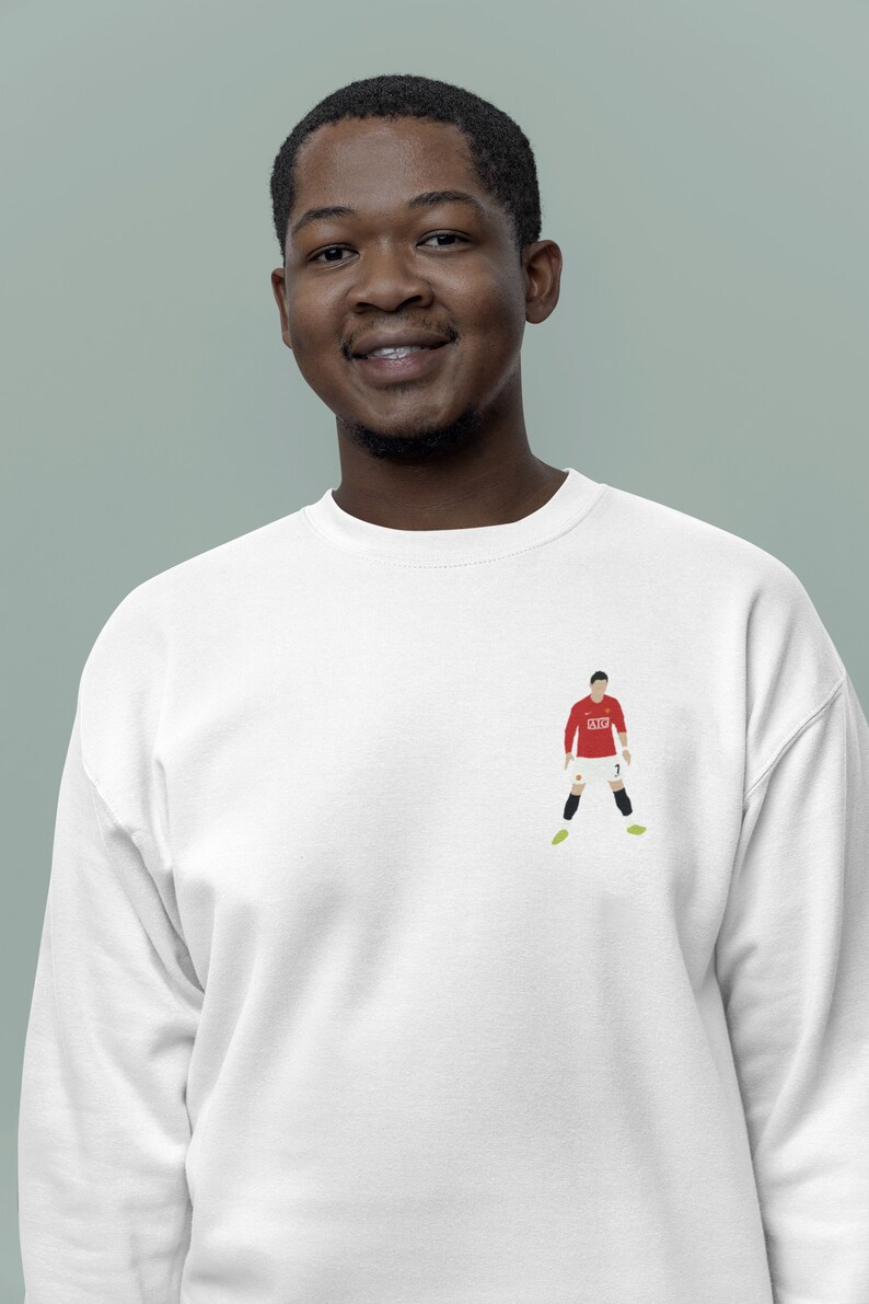 May include: A white sweatshirt with a red and white illustration of a soccer player wearing a red jersey with the number 7 on it. The player is wearing black shorts and yellow cleats.