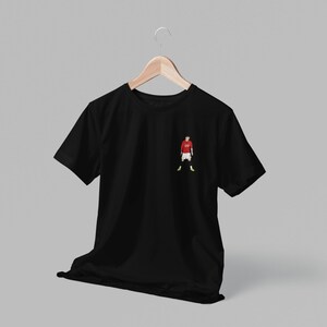 May include: Black t-shirt with a small embroidered image of a soccer player wearing a red jersey, white shorts, and yellow socks.