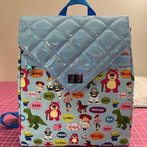 May include: A light blue quilted backpack with a flap closure. The backpack is decorated with a repeating pattern of Toy Story characters, including Woody, Buzz Lightyear, Jessie, and Lotso. The flap is iridescent and has a silver clasp.