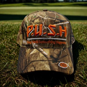 May include: Camouflage baseball cap with the word "PUSH" embroidered in orange. The text "Pray Until Something Happens" is embroidered below the word "PUSH". A small orange fish is embroidered on the side of the cap.