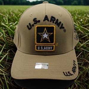 May include: Tan baseball cap featuring the U.S. Army logo. The logo includes a black and gold patch with a white star. The words "U.S. ARMY" are embroidered in black on the front and side of the cap. The cap is set against a background of green grass.