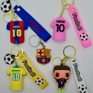 May include: Five different keychains featuring soccer players and team logos. The keychains are made of colorful rubber and feature the names "Messi" and "Neymar Jr." along with the team logo "FCB".