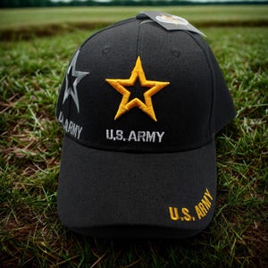 May include: Black baseball cap with a gold star and the words "U.S. ARMY" embroidered in white. The cap also features a silver star and the words "ARMY" in silver. The cap is sitting on green grass.